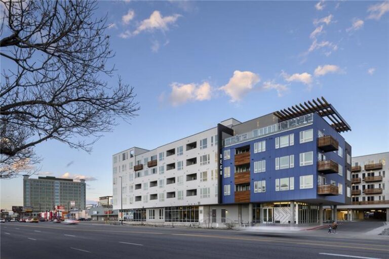 Deco Apartments en Denver, un edificio moderno de mediana altura con un exterior azul y blanco, balcones con detalles de madera y ventanas de piso a techo a nivel de la calle.